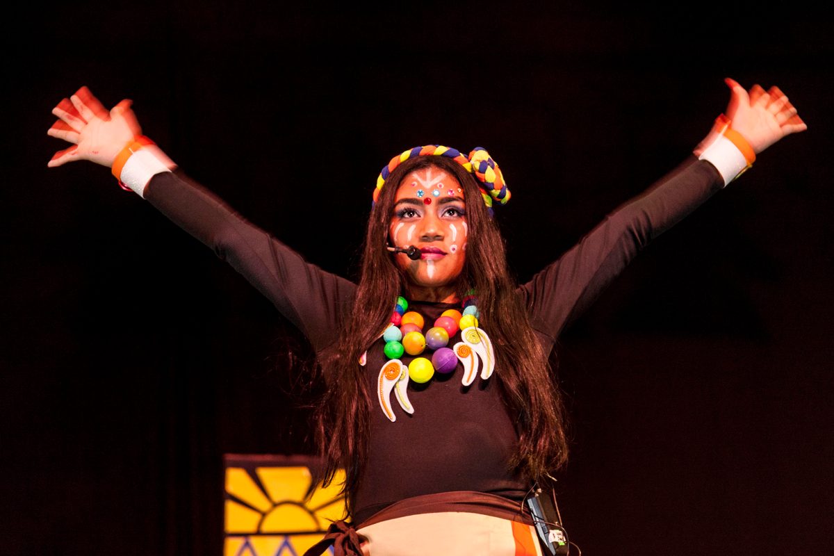 A young performer in a colourful necklace and feathered headband opens her arms on stage