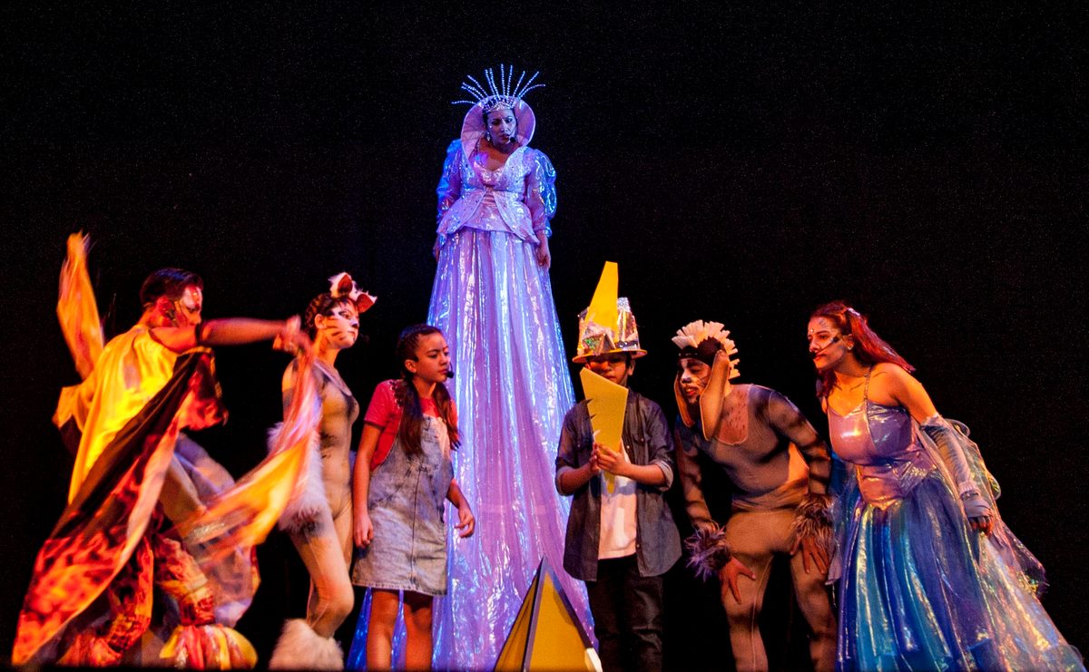 A character on stilts in a long gown, surrounded by the cast