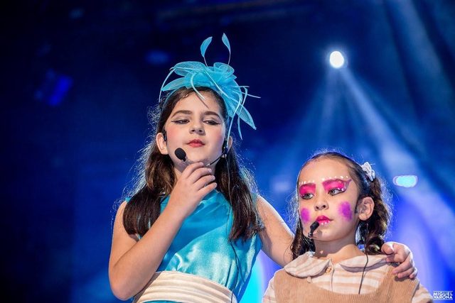 Dos niñas cantan con micrófono, con tocados de plumas, bajo luces azules