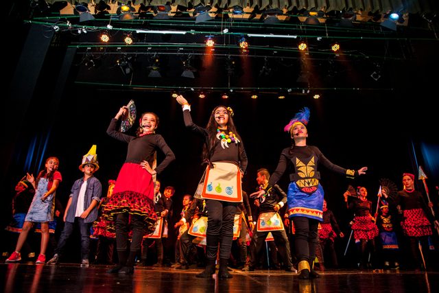Tres jóvenes con vestuario de inspiración indígena bailan al frente del elenco
