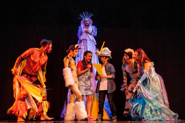 Bailarines con vestuario de colores frente a un personaje sobre zancos