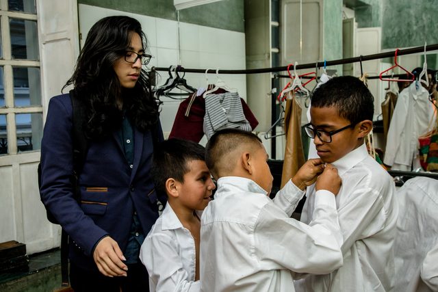 Niños se ayudan a vestirse entre bastidores, junto a una profesora