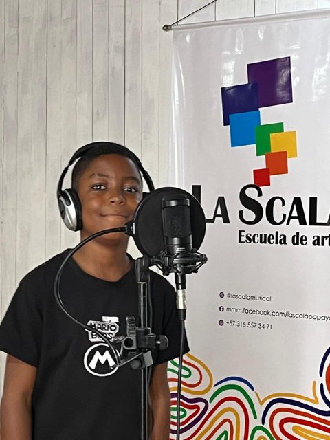 Un niño con audífonos canta frente a un micrófono, junto a un pendón de La Scala